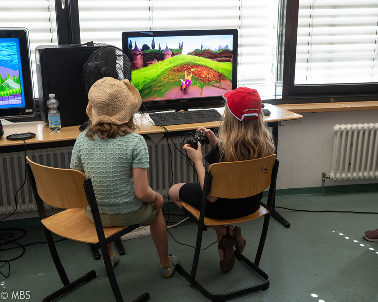 Zwei Kinder sitzen vor einem Monitor und spielen ein farbenfrohes Spiel mit einem Controller